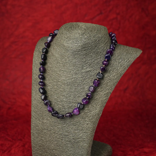 A beaded necklace made of irregular dark purple and black beads, displayed on a gray fabric jewelry bust against a textured red background.