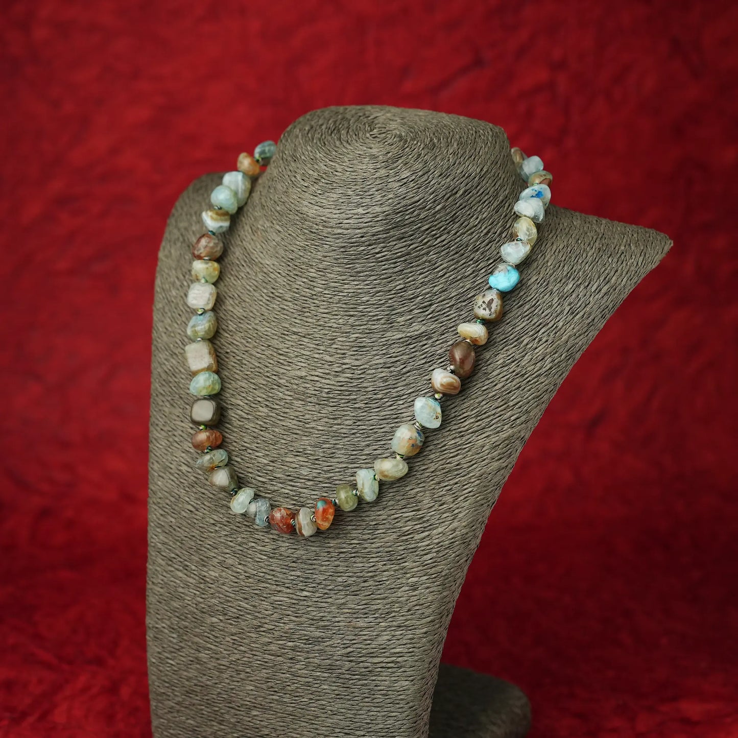 A beaded necklace made of irregular polished stone beads in earthy brown, cream, light blue, and green tones, displayed on a gray fabric jewelry bust against a textured red background.
