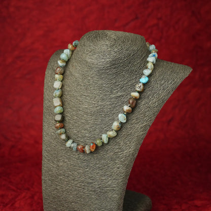 A beaded necklace made of irregular polished stone beads in earthy brown, cream, light blue, and green tones, displayed on a gray fabric jewelry bust against a textured red background.