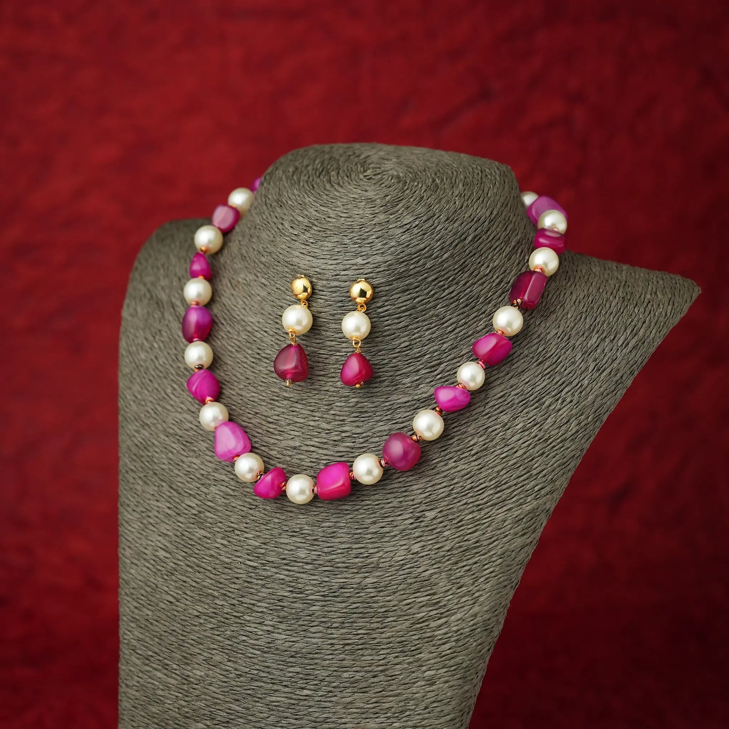 A jewelry set featuring a necklace and matching drop earrings displayed on a gray fabric mannequin bust against a textured red background. The necklace and earrings are made of white pearls and bright pink teardrop-shaped beads with gold-tone metal findings.