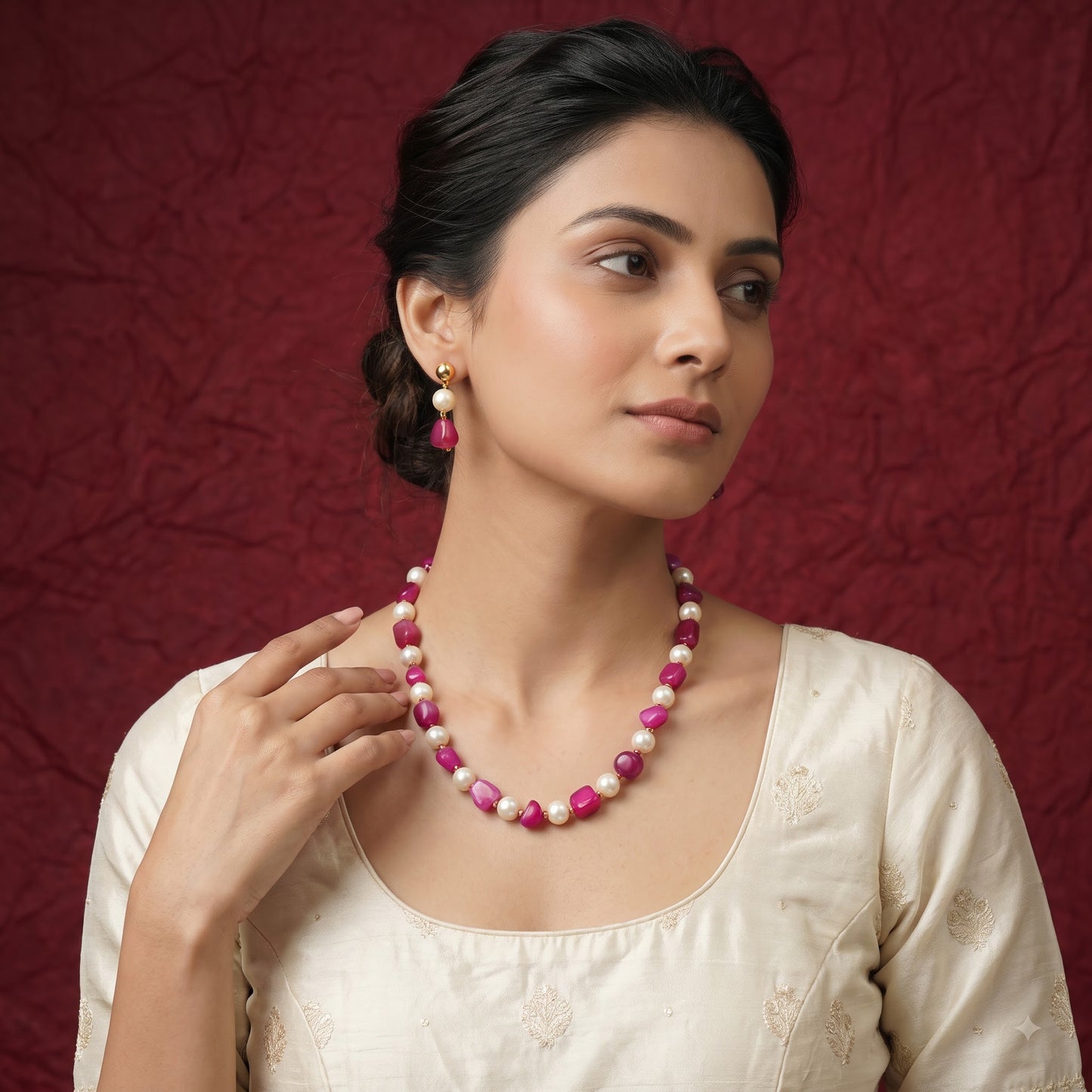 A woman wearing a pink and white beaded necklace with matching drop earrings, posed against a textured dark red background.