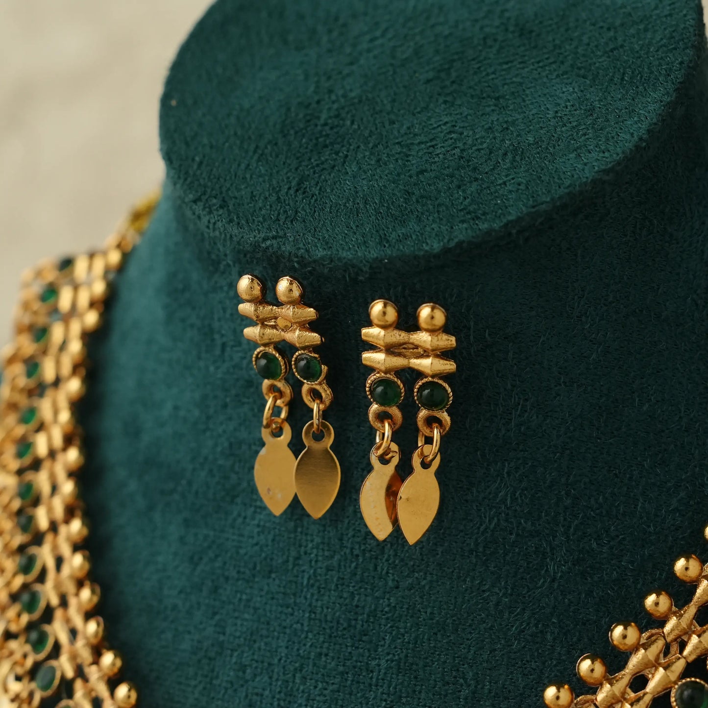 Gold-toned statement earrings with green stone accents and leaf-shaped dangling charms, displayed on a dark teal jewelry bust alongside a matching necklace.