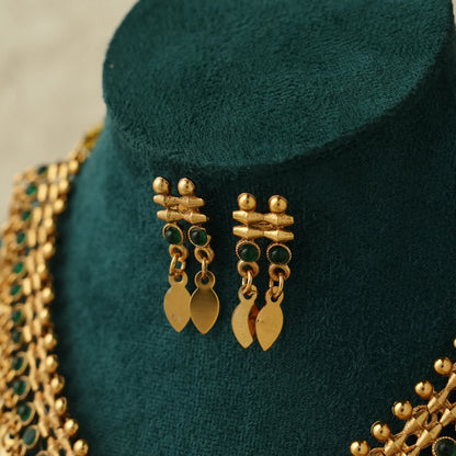 Gold-toned statement earrings with green stone accents and leaf-shaped dangling charms, displayed on a dark teal jewelry bust alongside a matching necklace.