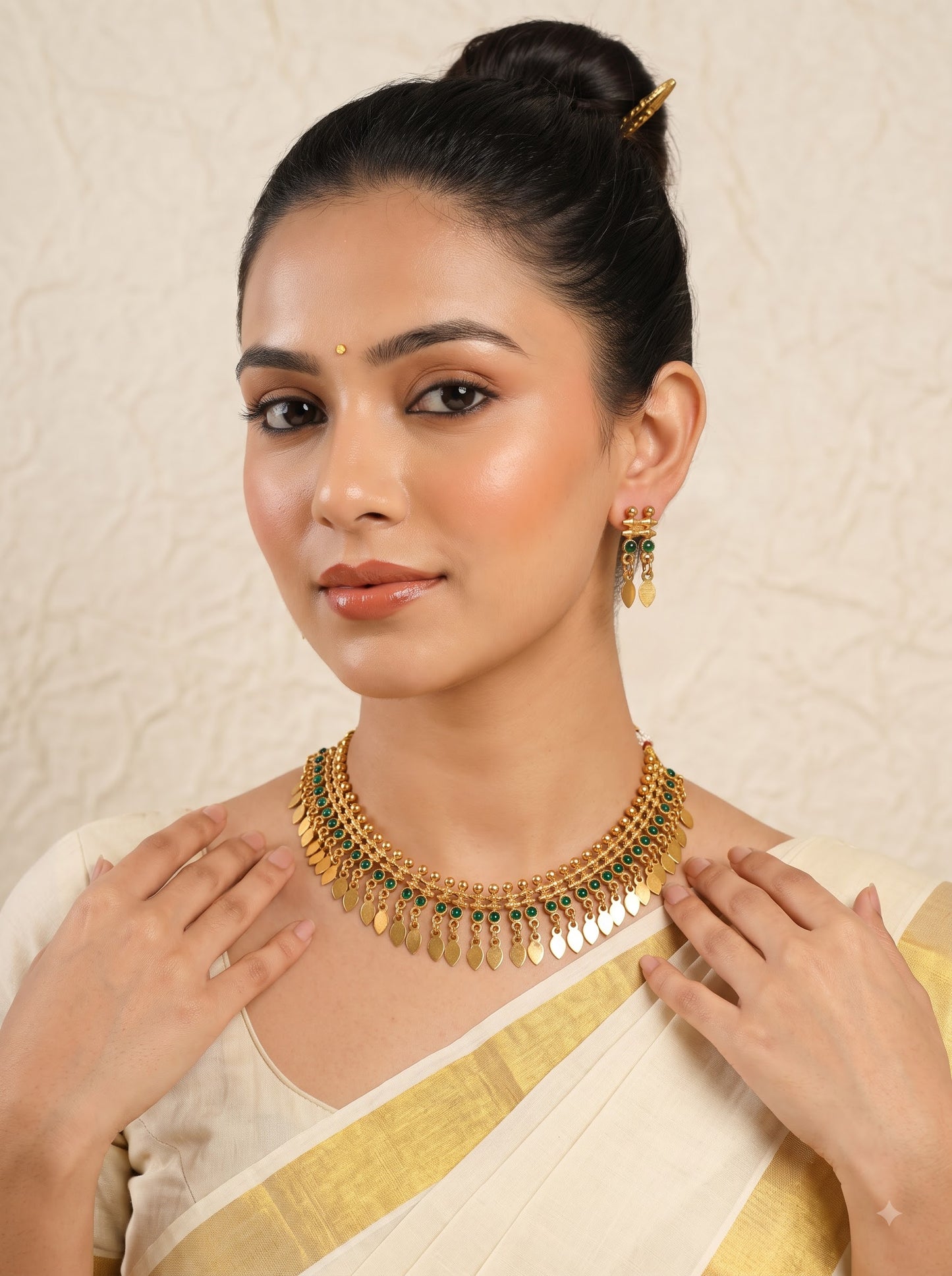 A woman wearing a gold-toned traditional necklace with green stone accents and matching dangling earrings, styled with a cream and gold sari against a textured beige background.