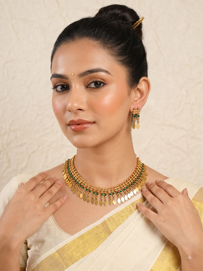 A woman wearing a gold-toned traditional necklace with green stone accents and matching dangling earrings, styled with a cream and gold sari against a textured beige background.