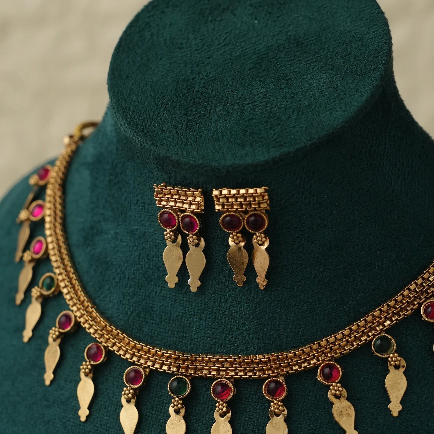 Gold-toned traditional jewelry set including a necklace and matching earrings displayed on a dark green velvet mannequin bust. The jewelry features gold-plated elements with red and green gemstone accents.