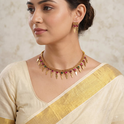 A woman wearing a gold-toned traditional necklace with leaf-shaped charms and small pink and green stone accents, paired with a cream and gold saree.