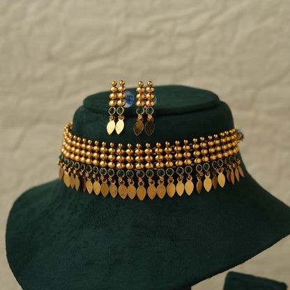 Gold-toned traditional jewelry set displayed on a dark green velvet jewelry bust, featuring a beaded necklace with hanging leaf-shaped coins, matching drop earrings, and a small blue stone accent.