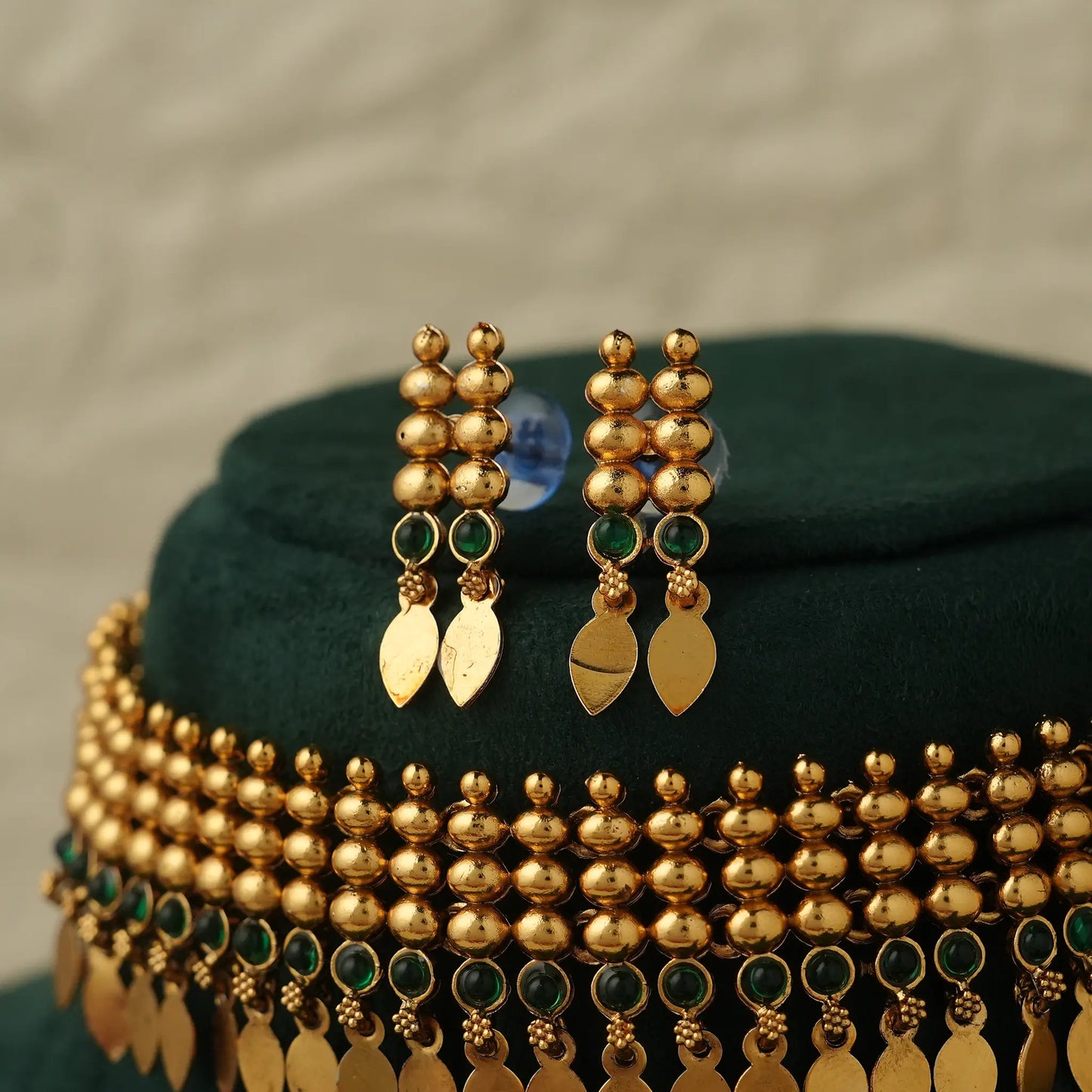 Gold-toned traditional Indian jewelry set featuring a choker necklace and matching drop earrings, decorated with gold beads, green stone accents, and dangling gold leaf-shaped charms, displayed on a dark green velvet stand.
