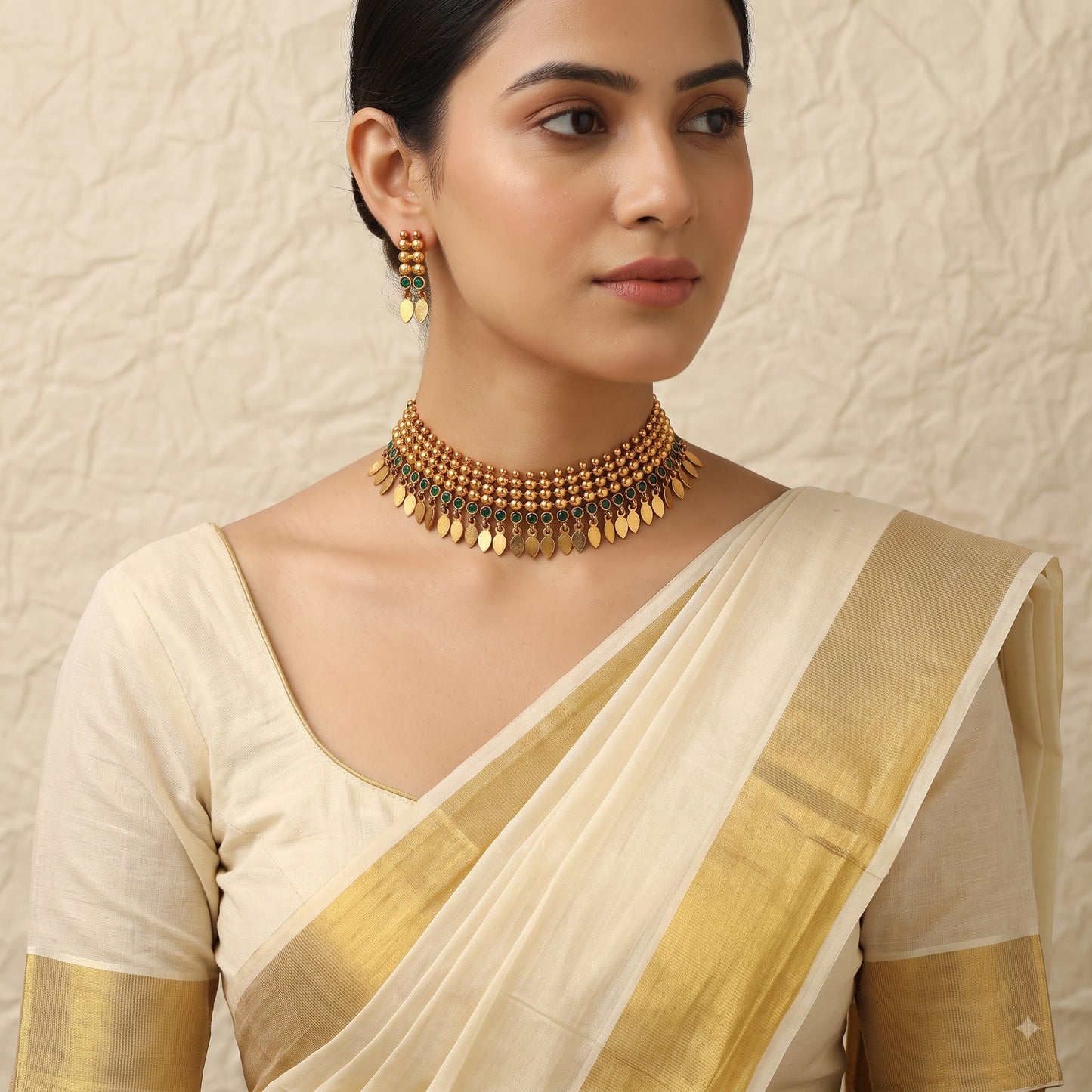 A woman wearing a cream-colored silk saree with a gold border and a matching gold coin choker necklace, set against a textured beige wall.