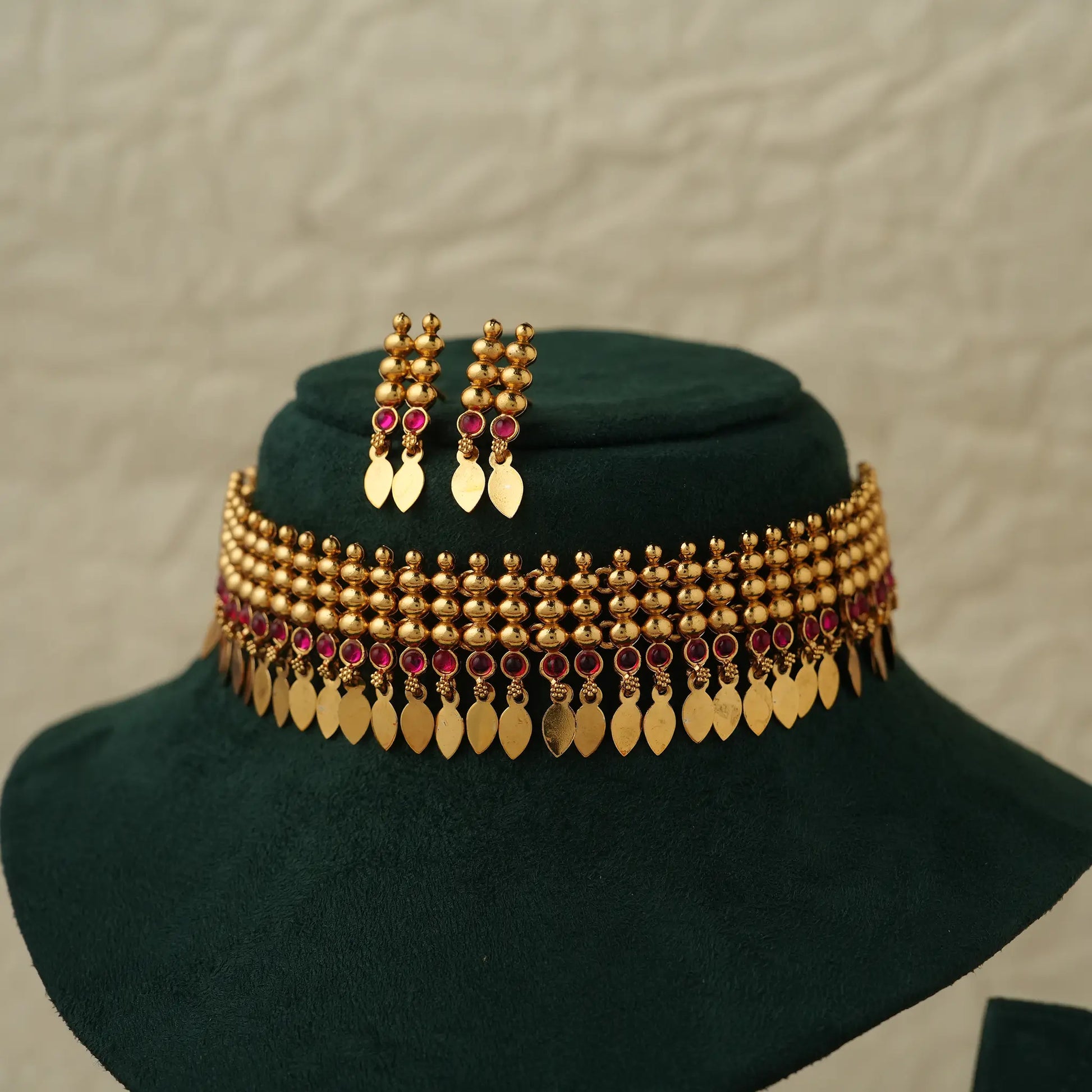 Gold-toned traditional Indian choker necklace with matching drop earrings, decorated with red stones and gold leaf-shaped pendants, displayed on a dark green velvet jewelry stand.
