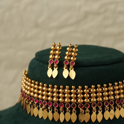 Gold-toned traditional Indian jewelry set featuring a choker necklace and matching drop earrings, decorated with gold beads, leaf-shaped dangling elements, and small ruby pink gemstone accents, displayed on a dark green velvet stand.