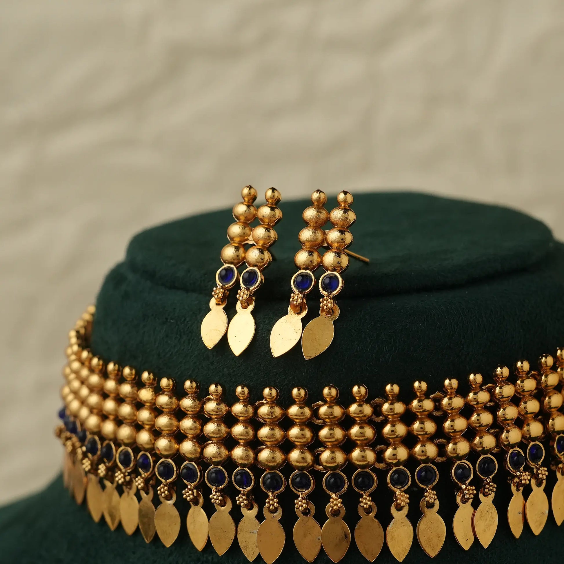 Gold-toned traditional jewelry set displayed on a dark green velvet stand, featuring a beaded necklace with dangling leaf-shaped charms and matching drop earrings with small blue stone accents.