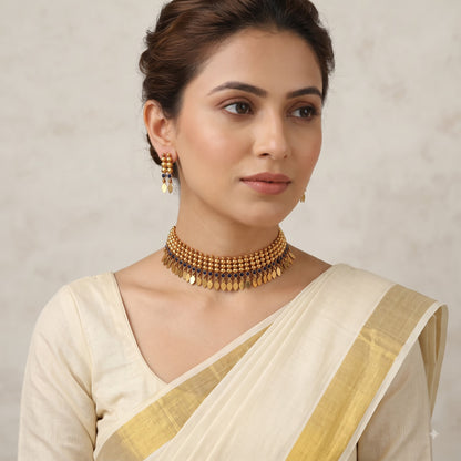A woman wearing a gold-toned traditional necklace with dangling coin-like charms and matching earrings, paired with a cream-colored sari with a gold border, against a soft neutral background.