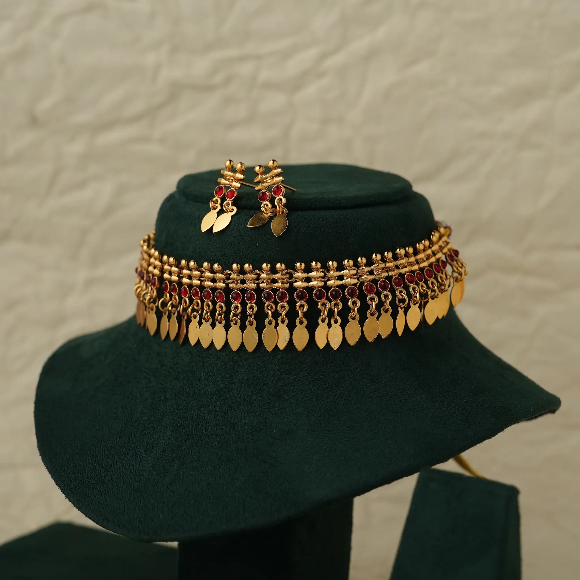 Gold-toned traditional Indian jewelry set including a wide chain necklace and matching earrings, decorated with gold leaf-shaped discs and red stones, displayed on a dark green velvet hat stand.