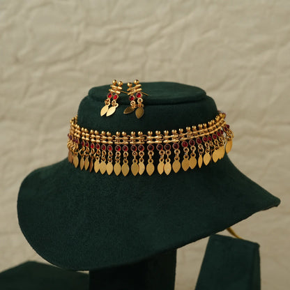 Gold-toned traditional Indian jewelry set including a wide chain necklace and matching earrings, decorated with gold leaf-shaped discs and red stones, displayed on a dark green velvet hat stand.