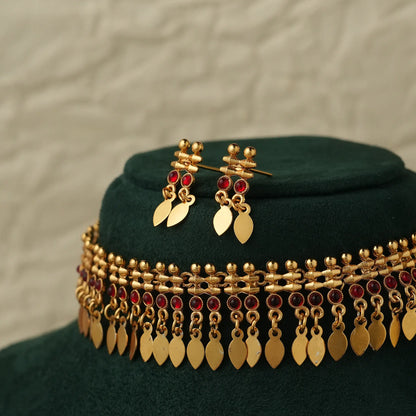 Gold-toned traditional jewelry set featuring a choker necklace and matching drop earrings, decorated with gold leaf-shaped pendants and small red stones, displayed on a dark green velvet stand.