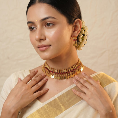 A woman wearing a traditional Indian gold-toned choker necklace with dangling disc elements and matching earrings, styled with a cream-colored saree against a textured beige background.