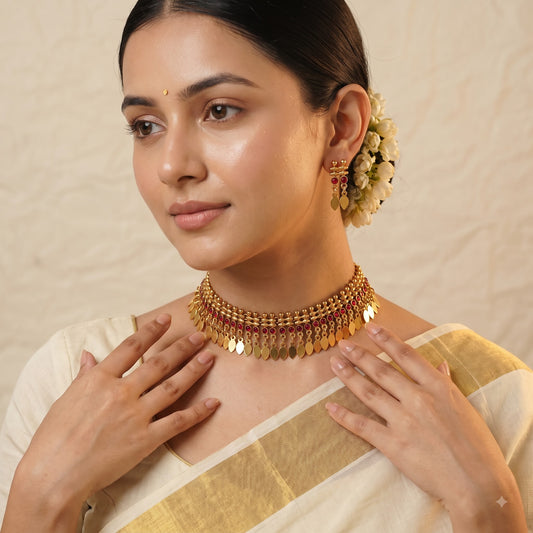 A woman wearing a traditional Indian gold-toned choker necklace with dangling disc elements and matching earrings, styled with a cream-colored saree against a textured beige background.