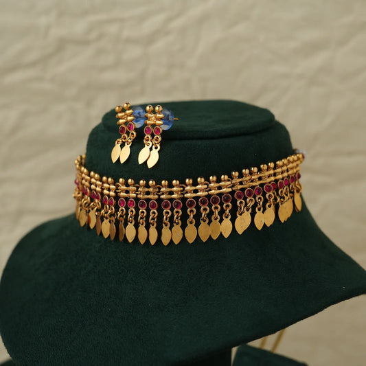 A gold-toned traditional Indian jewelry set displayed on a dark green velvet stand. The set includes a multi-strand chain necklace with gold leaf-shaped dangling charms and deep pink faceted beads, along with matching drop earrings.