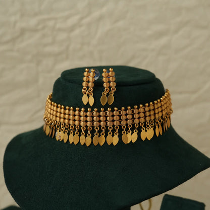 Gold-toned traditional Indian jewelry set featuring a multi-strand beaded necklace with dangling leaf-shaped coins, matching drop earrings, and a dark green velvet display stand against a textured light beige background.