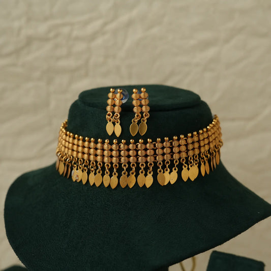 Gold-toned traditional Indian jewelry set featuring a multi-strand beaded necklace with dangling leaf-shaped coins, matching drop earrings, and a dark green velvet display stand against a textured light beige background.