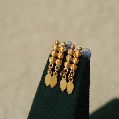 A pair of gold-toned drop earrings with stacked textured beads, small round beads, and two leaf-shaped dangles, displayed on a dark green jewelry stand against a soft beige background.