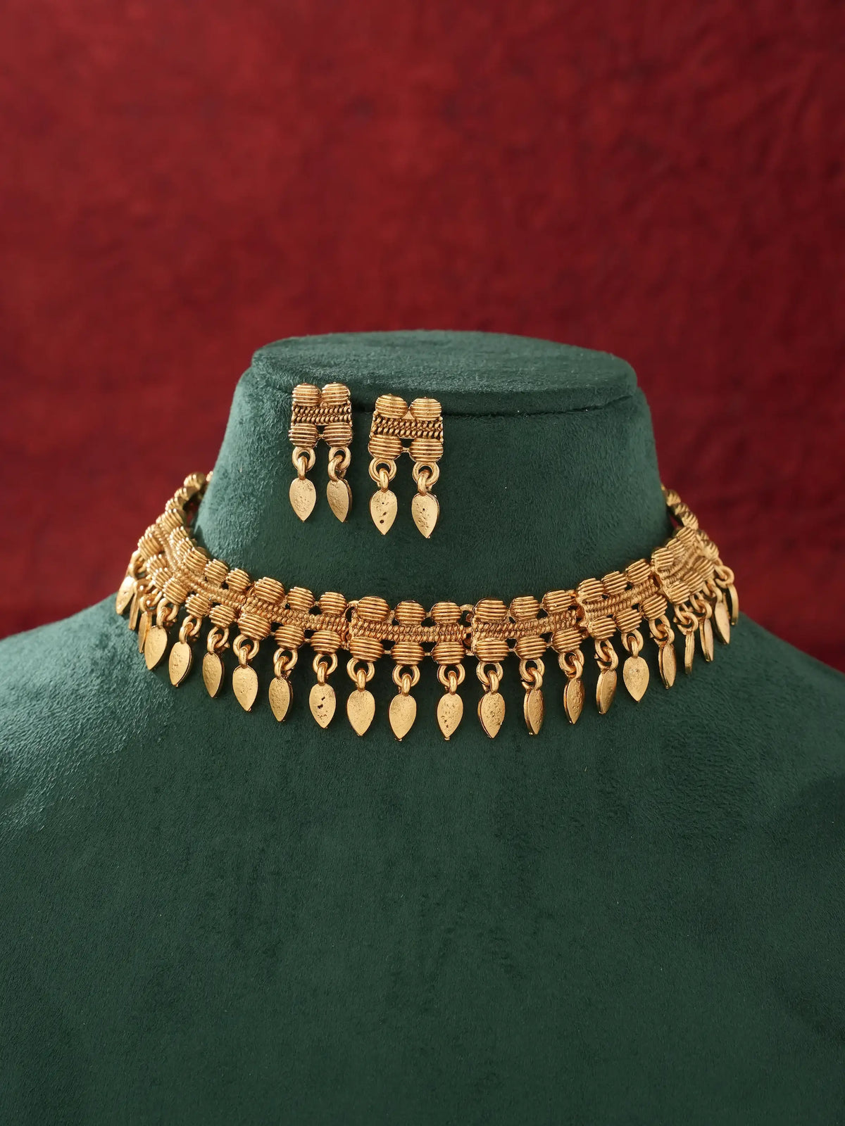 Gold-toned traditional necklace and matching drop earrings displayed on a dark green velvet jewelry bust against a textured red background.
