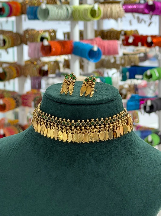 A gold coin choker necklace and matching gold earrings displayed on a dark green velvet jewelry bust. The background shows a blurred shop display filled with colorful bangles.