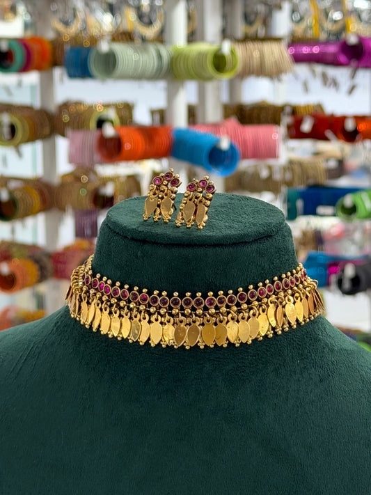 Gold-toned traditional Indian jewelry set featuring a coin choker necklace and matching earrings with pink stones, displayed on a dark green velvet mannequin bust.