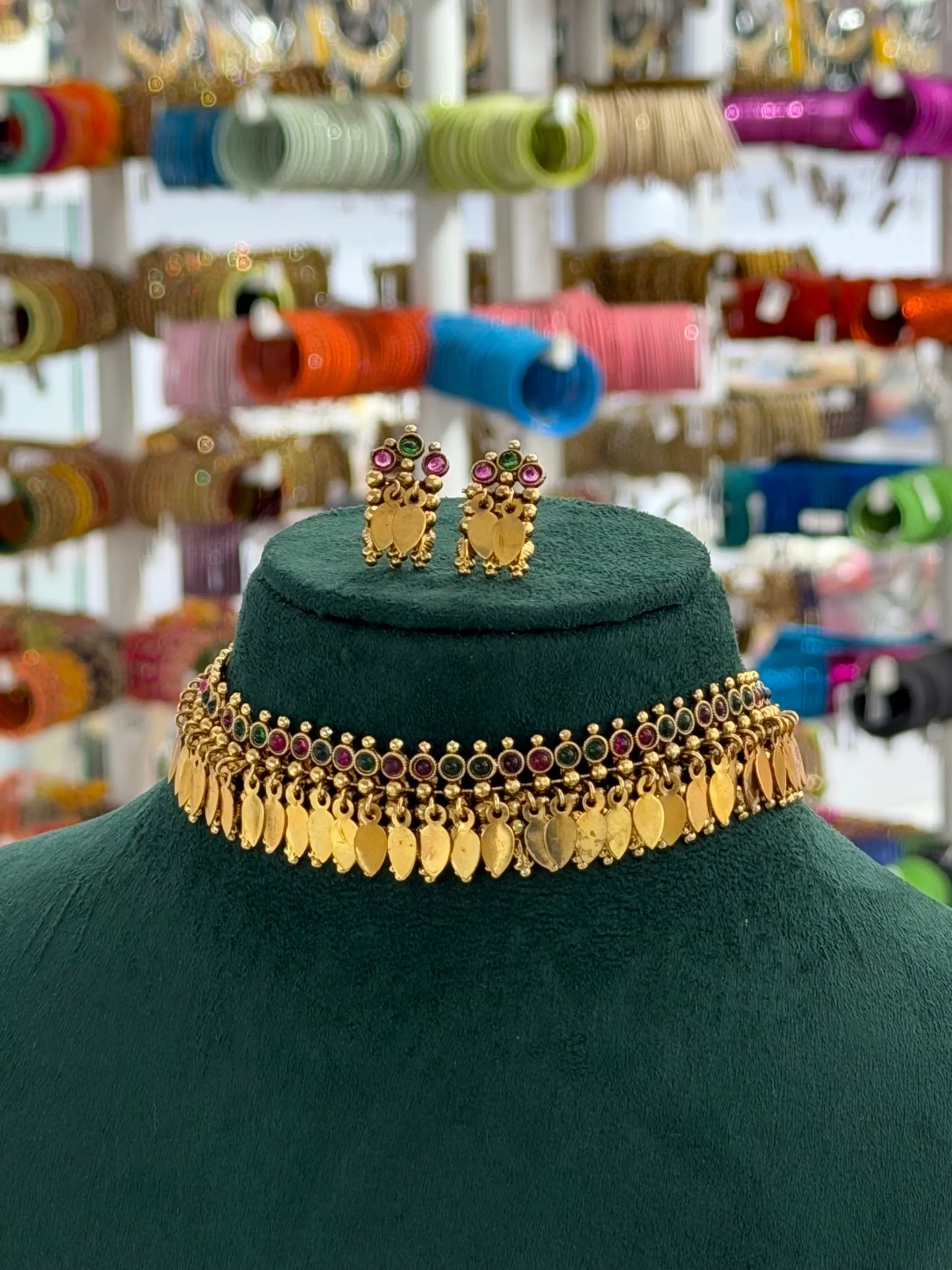 A gold-toned Indian-style choker necklace with dangling leaf-shaped coins and small red and green stone accents, displayed on a dark green velvet mannequin bust. Matching owl-shaped stud earrings are placed on top of the bust.