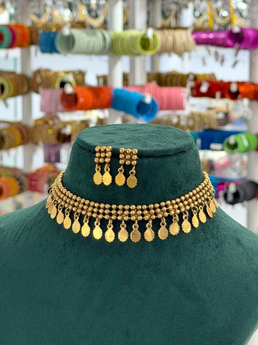 A gold-toned beaded necklace with dangling coin charms and matching drop earrings displayed on a dark green velvet mannequin bust, with a blurred jewelry shop background filled with colorful bangles.