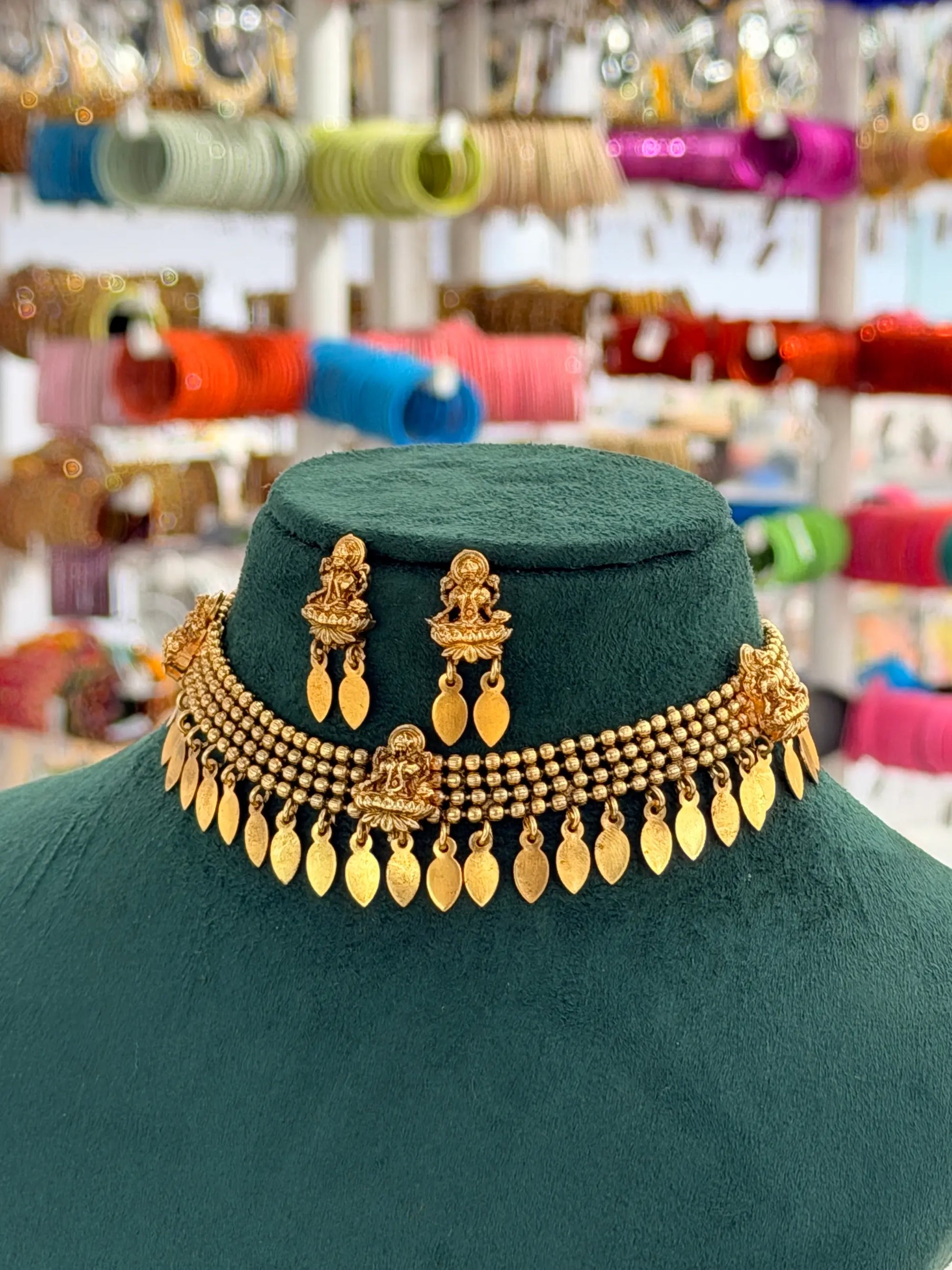 A gold-toned traditional Indian jewelry set displayed on a dark green velvet mannequin bust, featuring a multi-strand beaded necklace with hanging teardrop charms and matching drop earrings, with a blurred jewelry shop background.