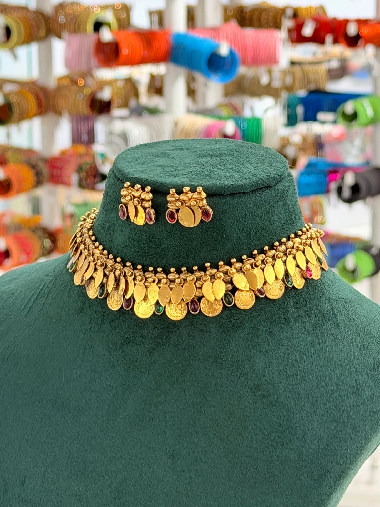 A gold coin-style choker necklace with dangling round coins and small red and green stone accents, displayed on a dark green velvet jewelry bust. The background shows a blurred shop interior with colorful bangles hanging on racks.