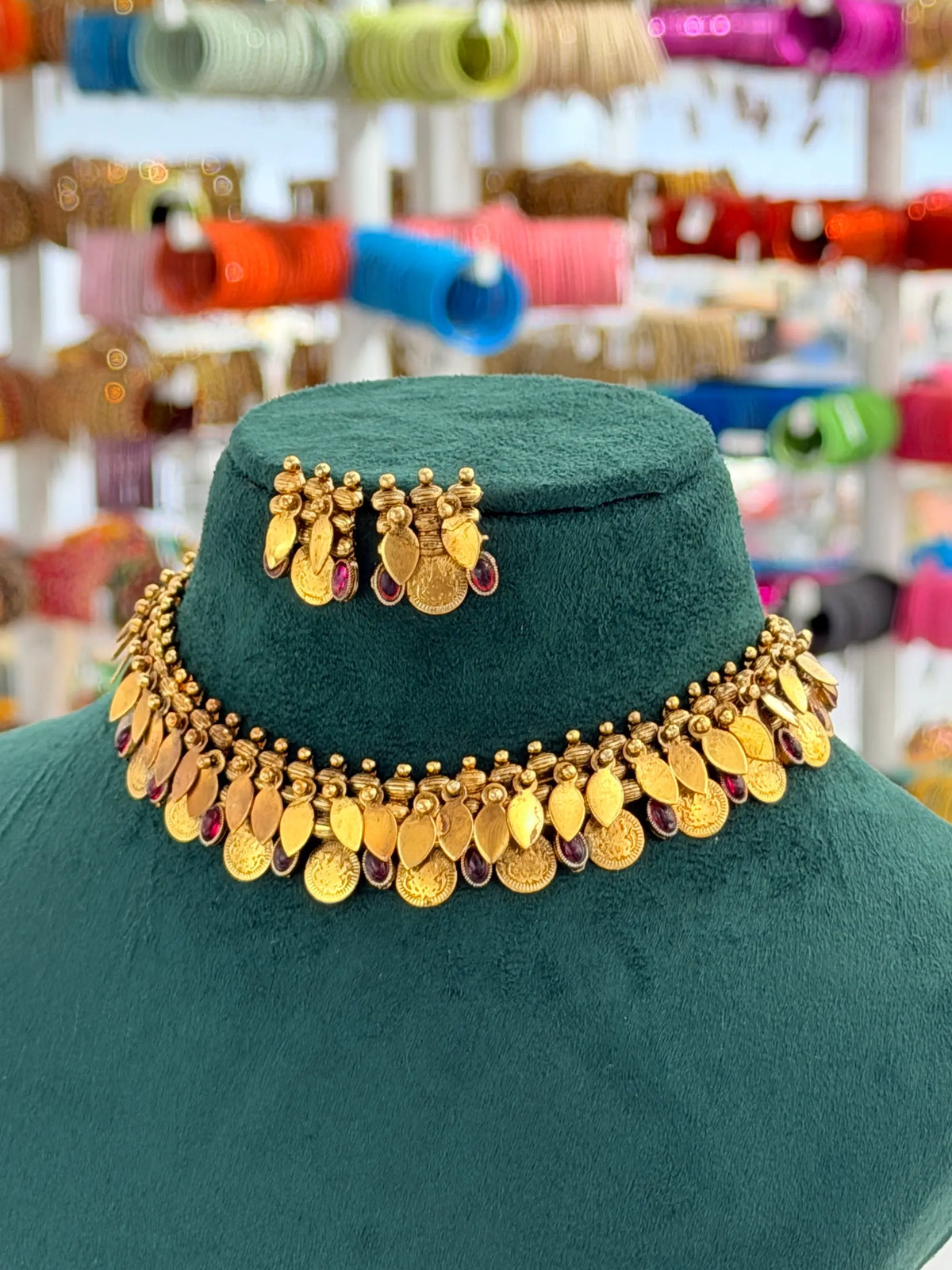 A gold coin-style necklace and matching earrings displayed on a dark green velvet jewelry bust, with a blurred shop background showing colorful spools of thread.