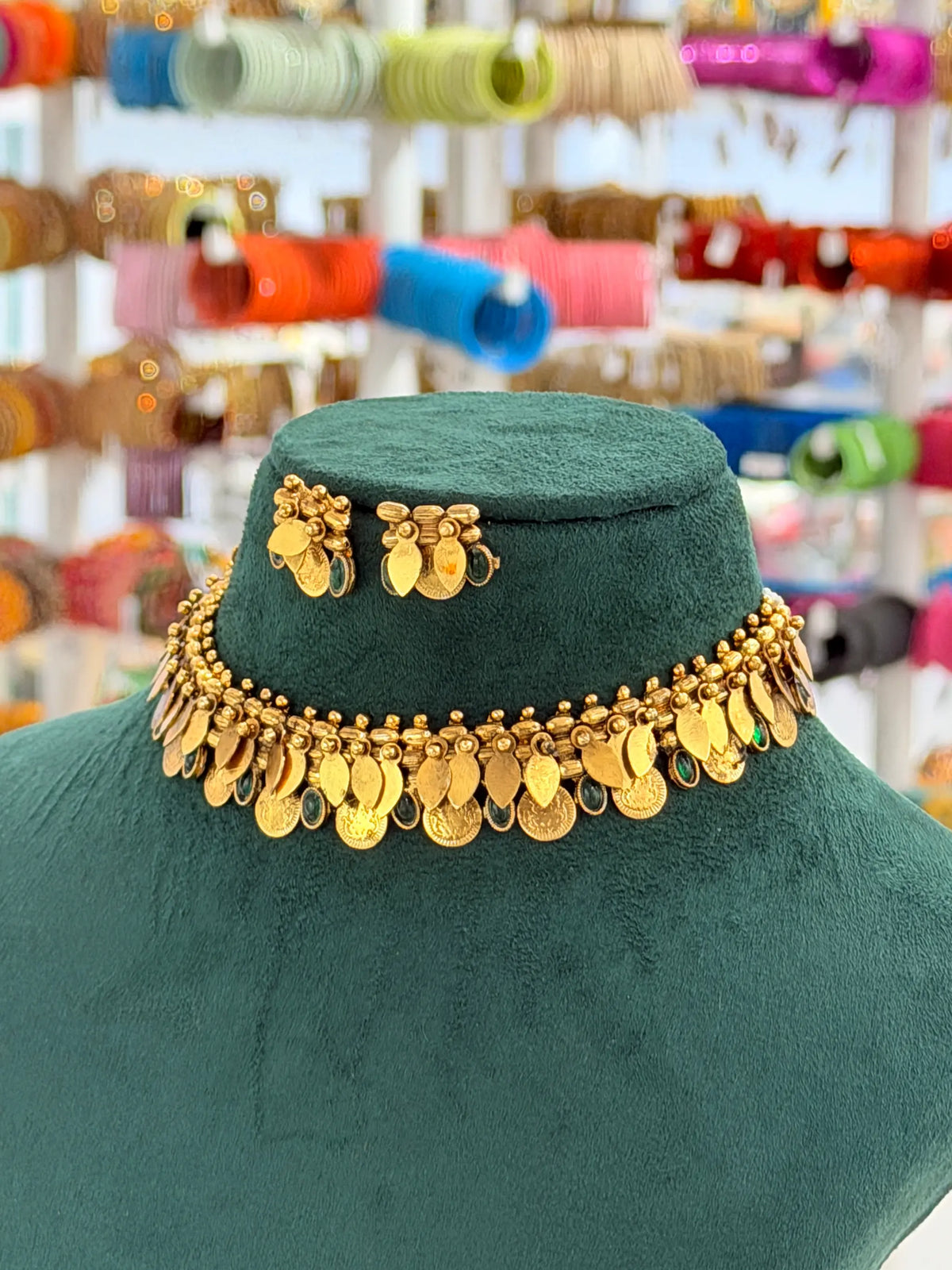A gold coin-style choker necklace with matching butterfly earrings displayed on a dark green velvet mannequin bust, with a blurred jewelry store background.