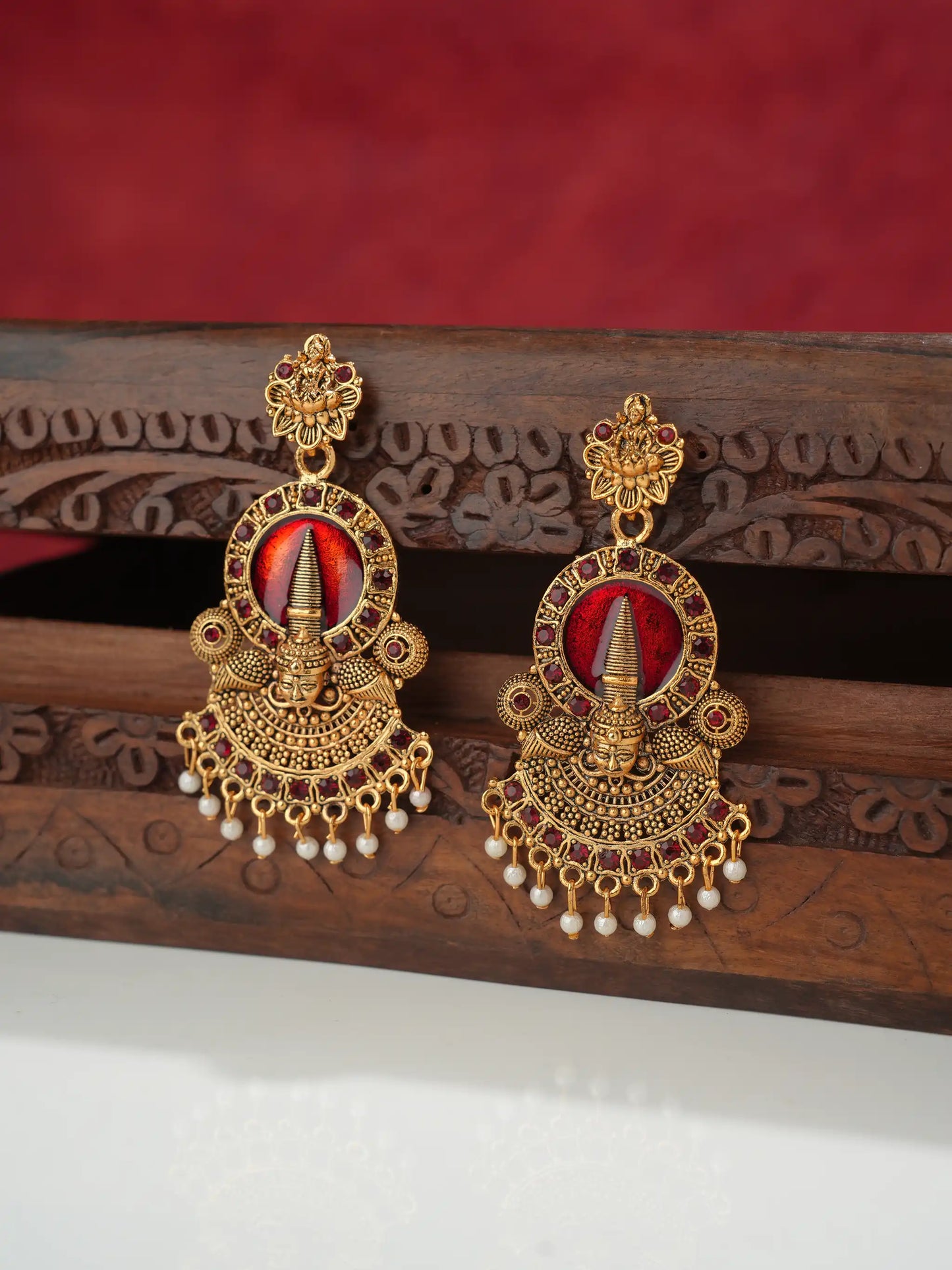 A pair of ornate gold-toned traditional Indian earrings with red and white stone inlays and dangling pearl-like beads, displayed on a carved wooden stand against a red background.