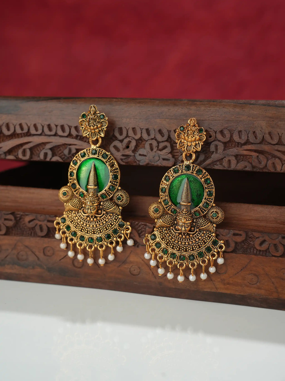 A pair of ornate gold-toned traditional earrings with green stone centers and dangling pearl-like beads, displayed on a carved wooden stand against a red fabric background.