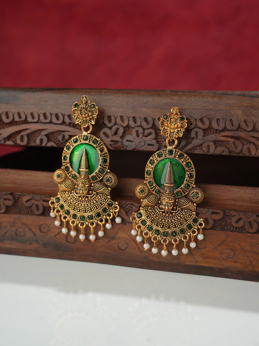 A pair of ornate gold-toned traditional earrings with green stone centers and dangling pearl-like beads, displayed on a carved wooden stand against a red fabric background.