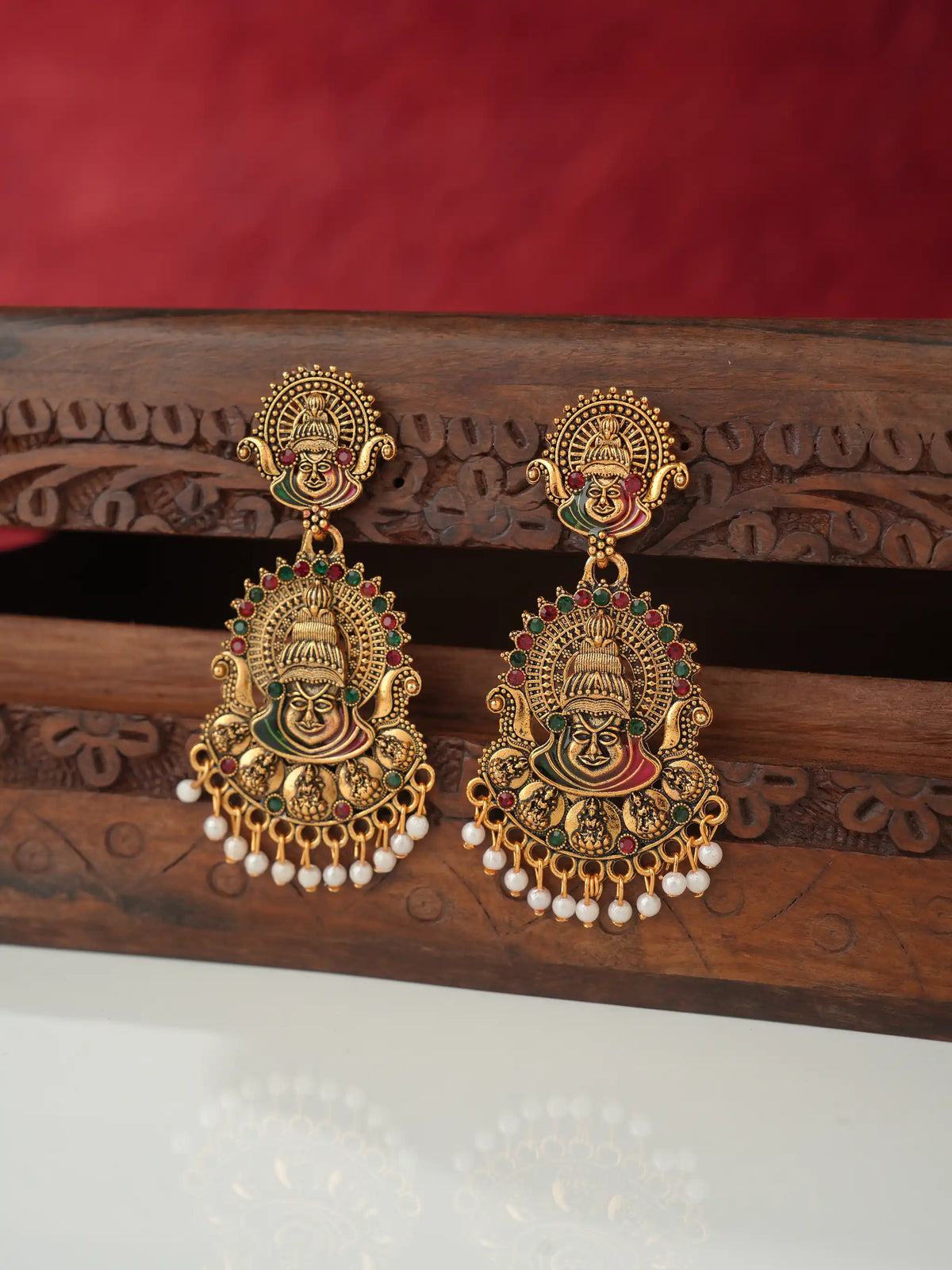 A pair of ornate gold-toned traditional Indian earrings with colorful enamel details, hanging from a carved wooden stand against a red background.