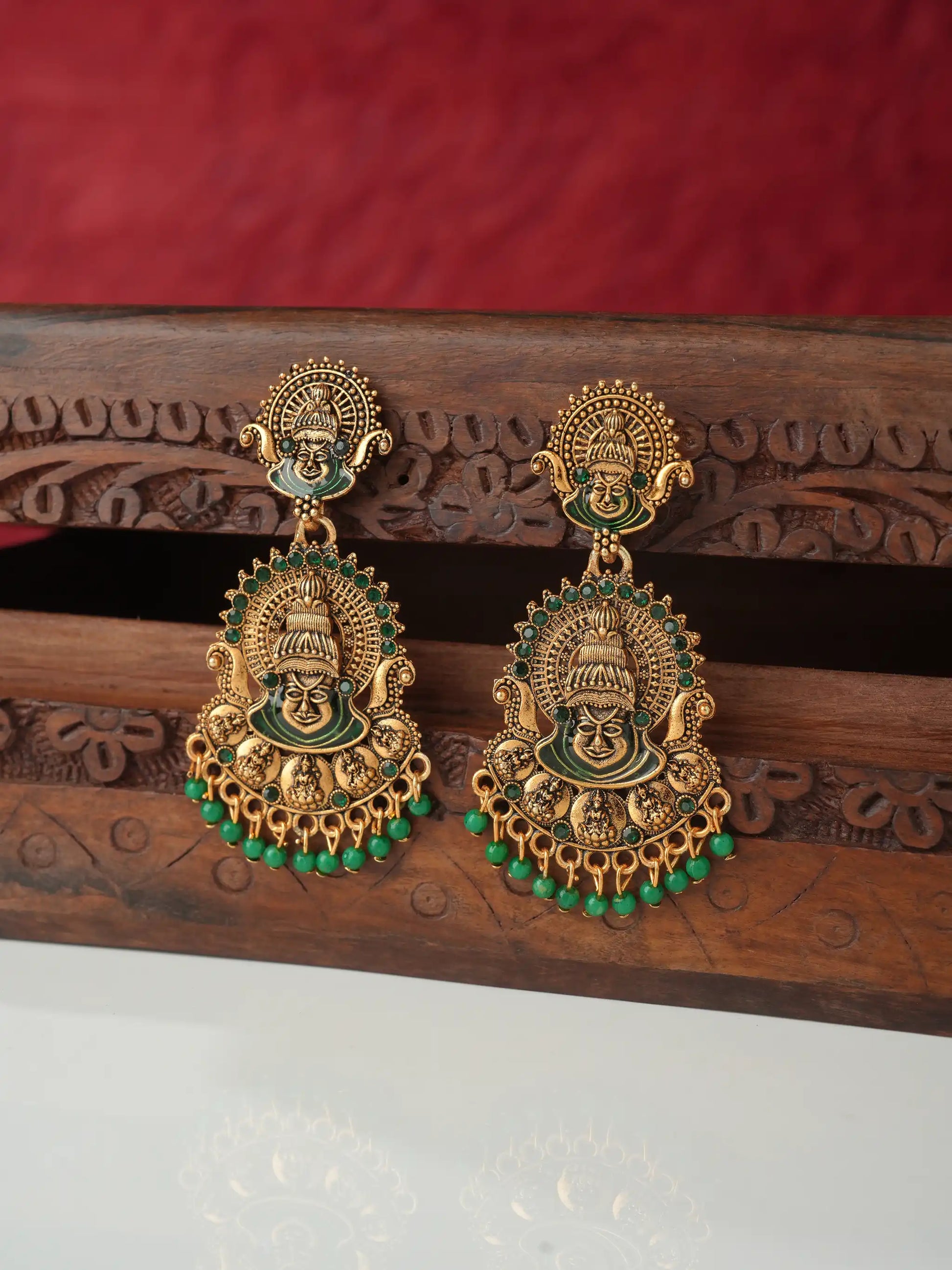 A pair of ornate gold-toned traditional earrings with green enamel and bead details, displayed on a carved wooden stand against a red background.