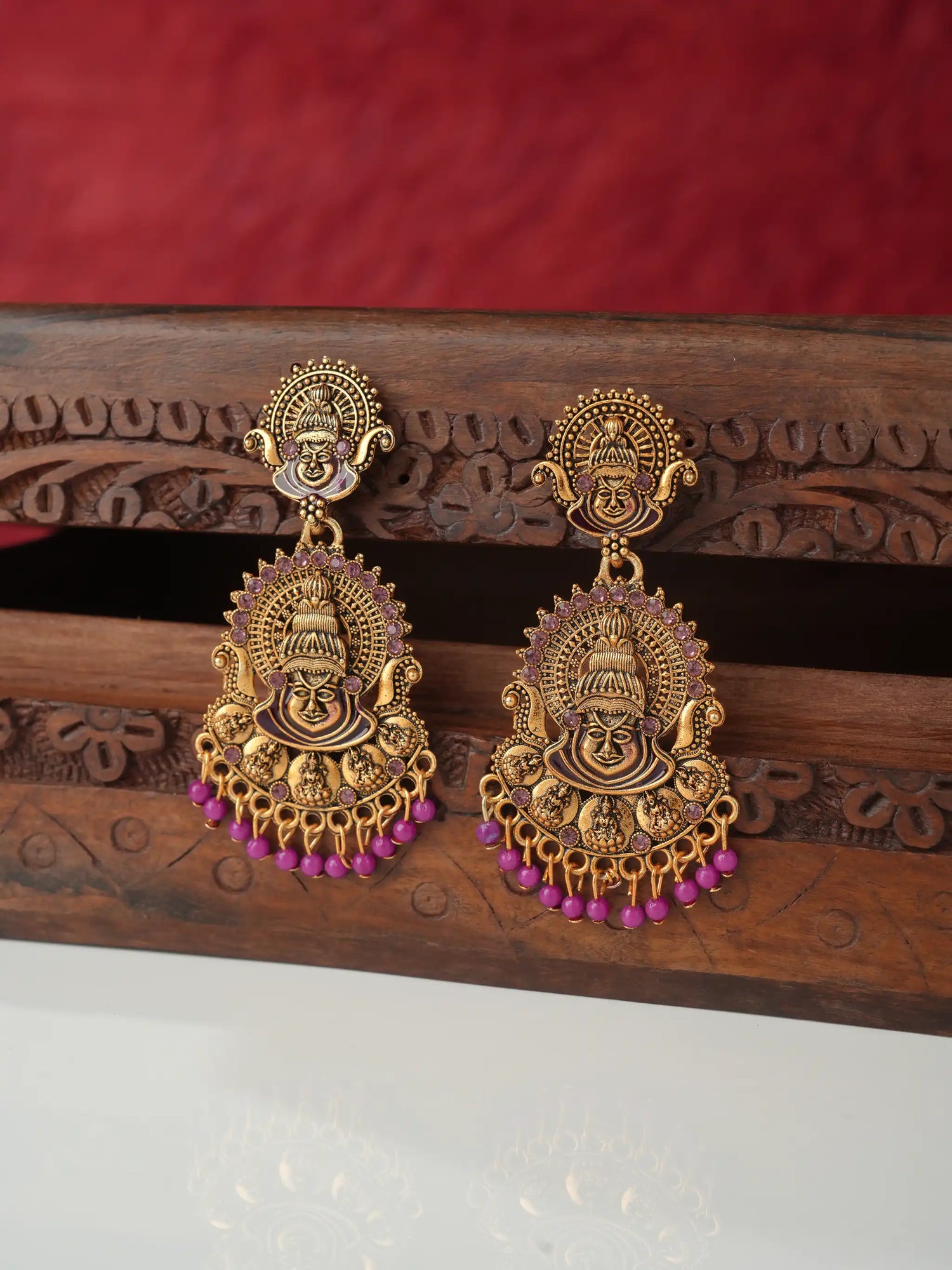 A pair of ornate gold-toned dangle earrings featuring detailed Hindu deity motifs and dangling pink beads, displayed on a carved wooden stand against a red textured background.