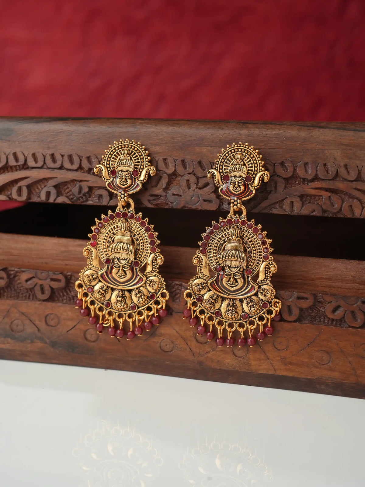 A pair of ornate gold-toned traditional Indian earrings featuring detailed deity motifs and dangling red bead accents, displayed on a carved wooden stand against a red fabric background.