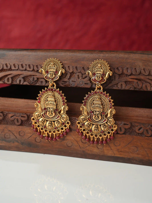 A pair of ornate gold-toned traditional Indian earrings featuring detailed deity motifs and dangling red bead accents, displayed on a carved wooden stand against a red fabric background.