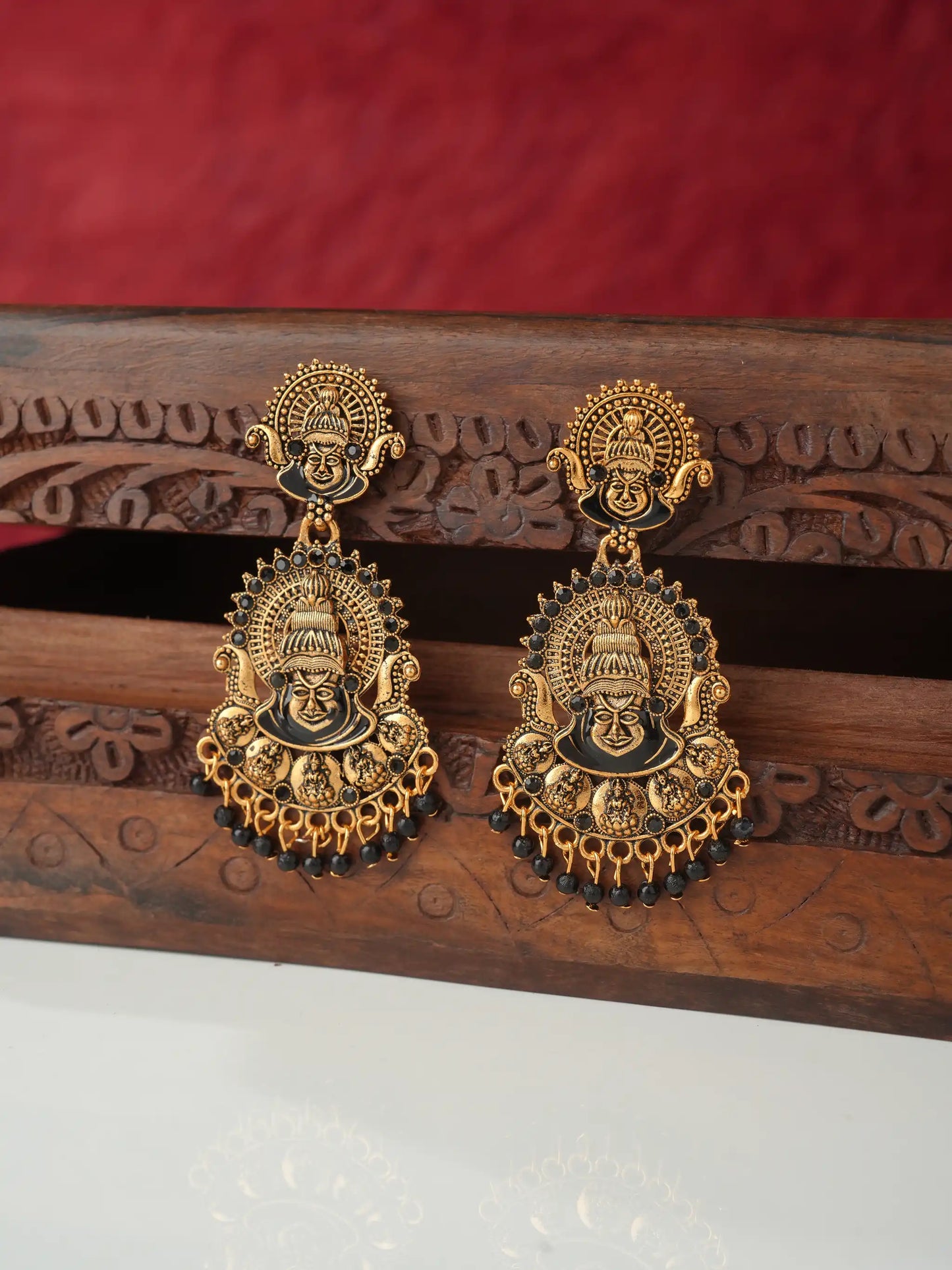 A pair of ornate gold-toned traditional Indian earrings featuring detailed deity motifs and dangling bead chains, displayed on a carved wooden stand against a red fabric background.
