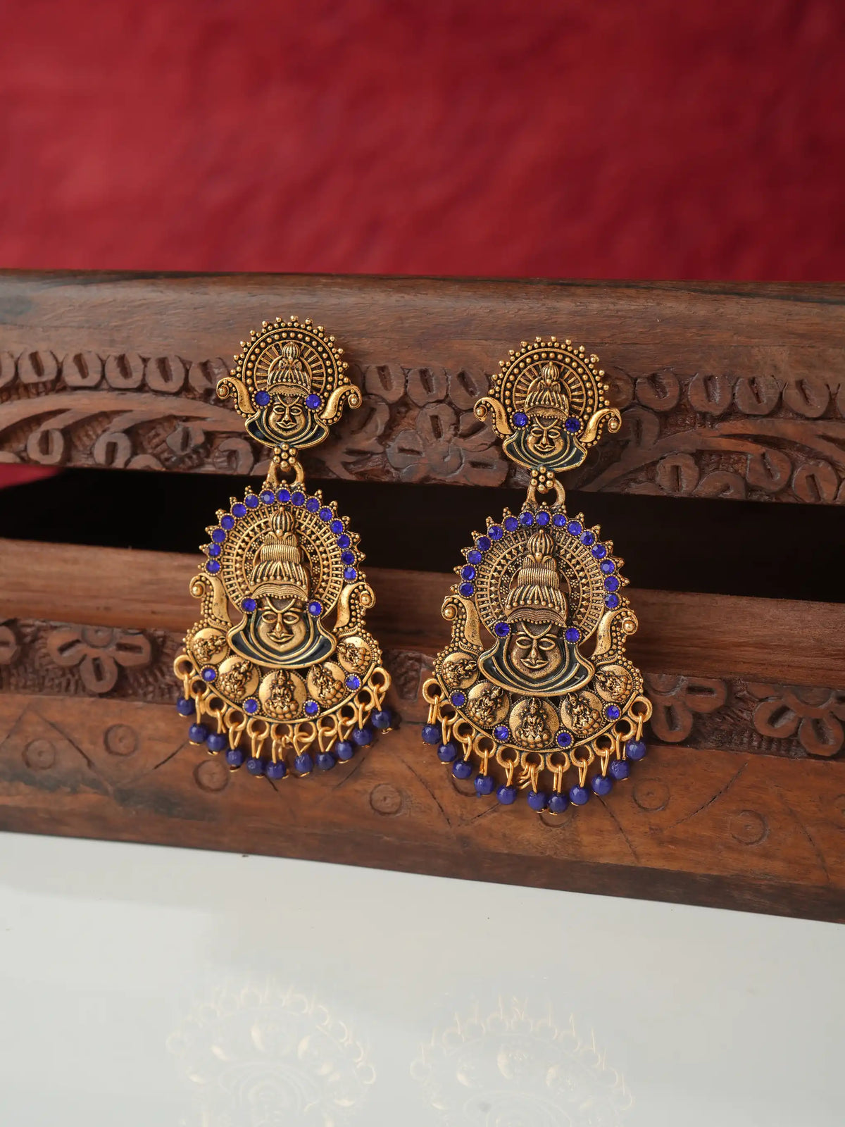 A pair of ornate gold-toned traditional earrings with blue stone accents, displayed on a carved wooden stand against a red fabric background.