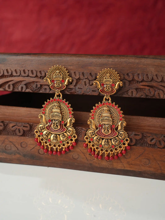 A pair of ornate gold-toned traditional earrings with red enamel detailing and dangling red bead drops, displayed on a carved wooden stand against a red fabric background.