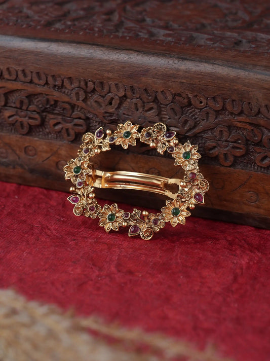 A gold-toned oval hair barrette decorated with small floral motifs and colorful gemstones, resting on a textured red fabric surface in front of a carved dark wood box.