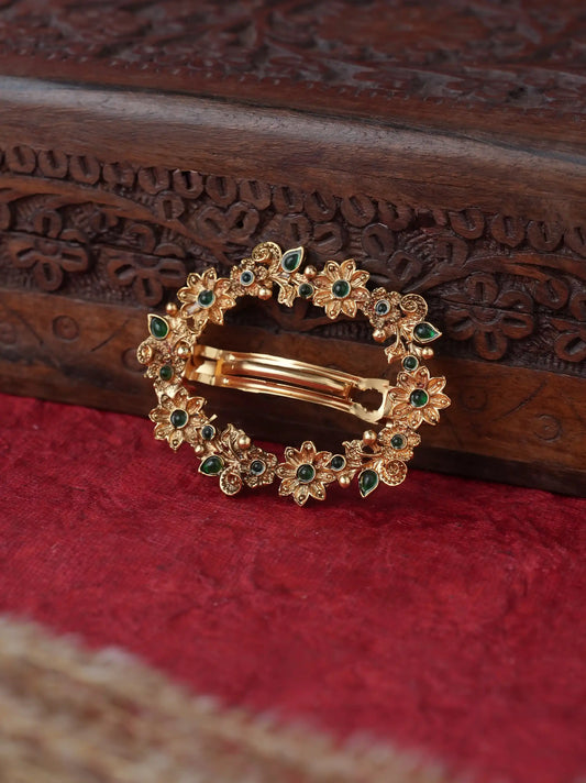A gold-toned hair claw clip decorated with small floral motifs and green stones, resting on a textured red fabric surface with a carved dark wood box in the background.