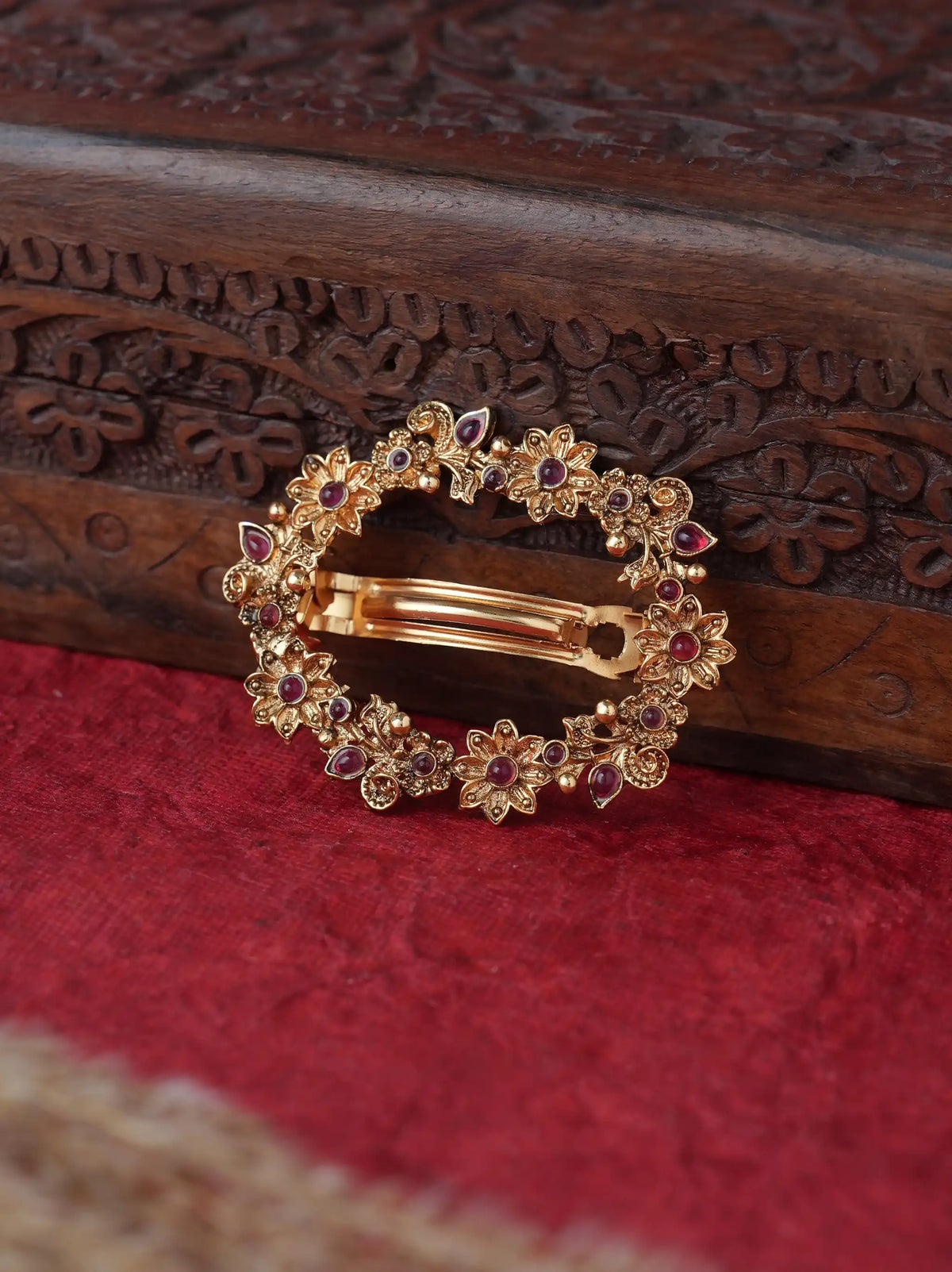 A gold-toned circular hair claw clip decorated with small floral motifs and red gemstones, resting on a textured red fabric surface next to a carved dark wood box.